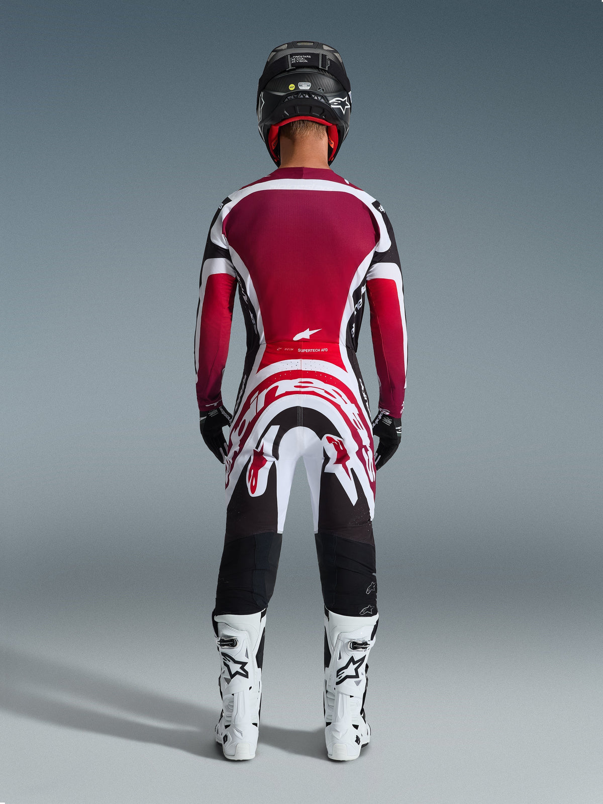 Piloto de motocross visto de costas a usar a camisola Alpinestars 2026 Supertech AFD Vista em preto, vermelho e branco, combinada com calças de motocross a condizer, capacete preto e botas brancas, conjunto completo de equipamento de corrida profissional para competição off-road