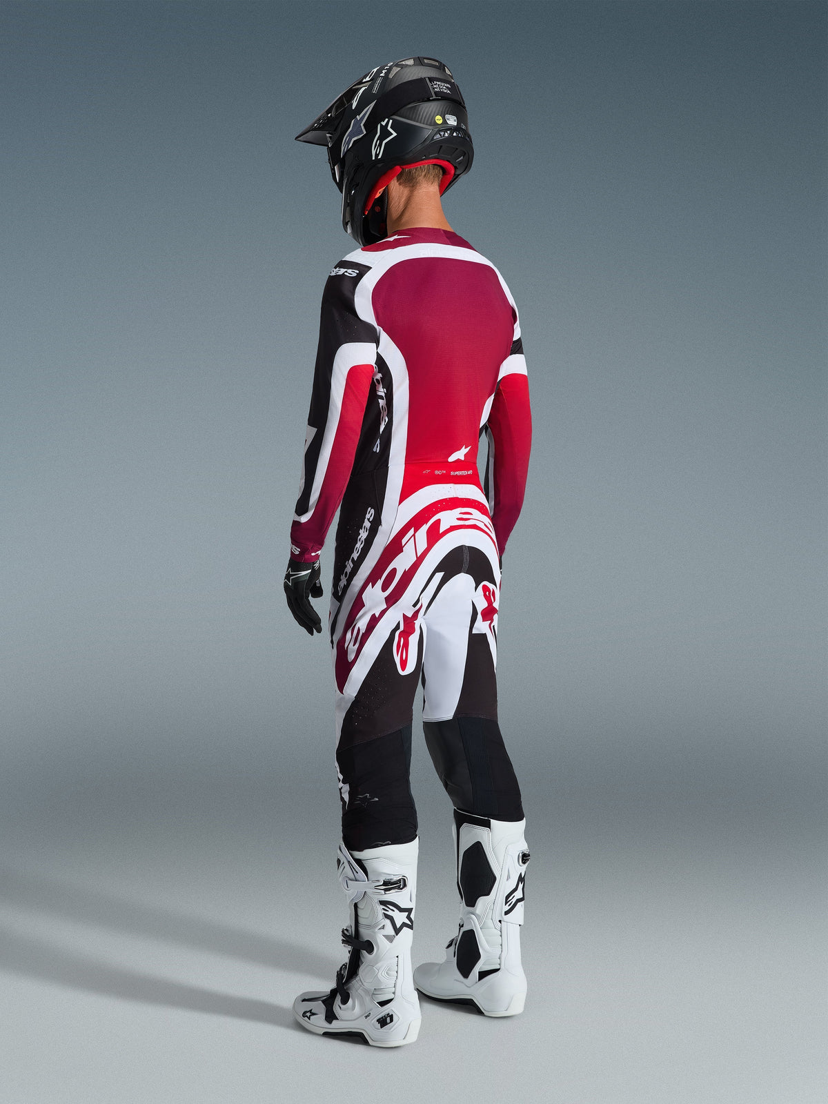 Piloto de motocross visto de costas a usar a camisola 2026 Supertech AFD Vista em preto, vermelho e branco, calças de motocross a condizer com a marca Alpinestars em destaque a branco, capacete preto e botas brancas e pretas, conjunto de equipamento profissional de corrida off-road