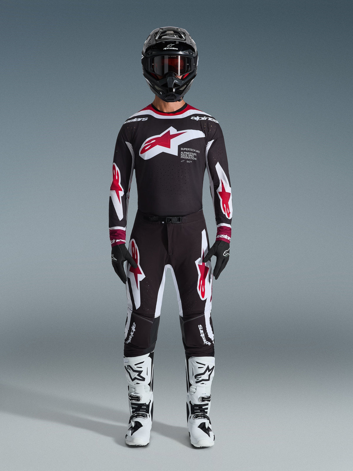 Piloto de motocross a usar a camisola Alpinestars 2026 Supertech AFD Vista, equipamento em preto, vermelho e branco, calças pretas a condizer com logótipos de estrela vermelhos, botas brancas e pretas, capacete preto com óculos, kit de competição off-road profissional
