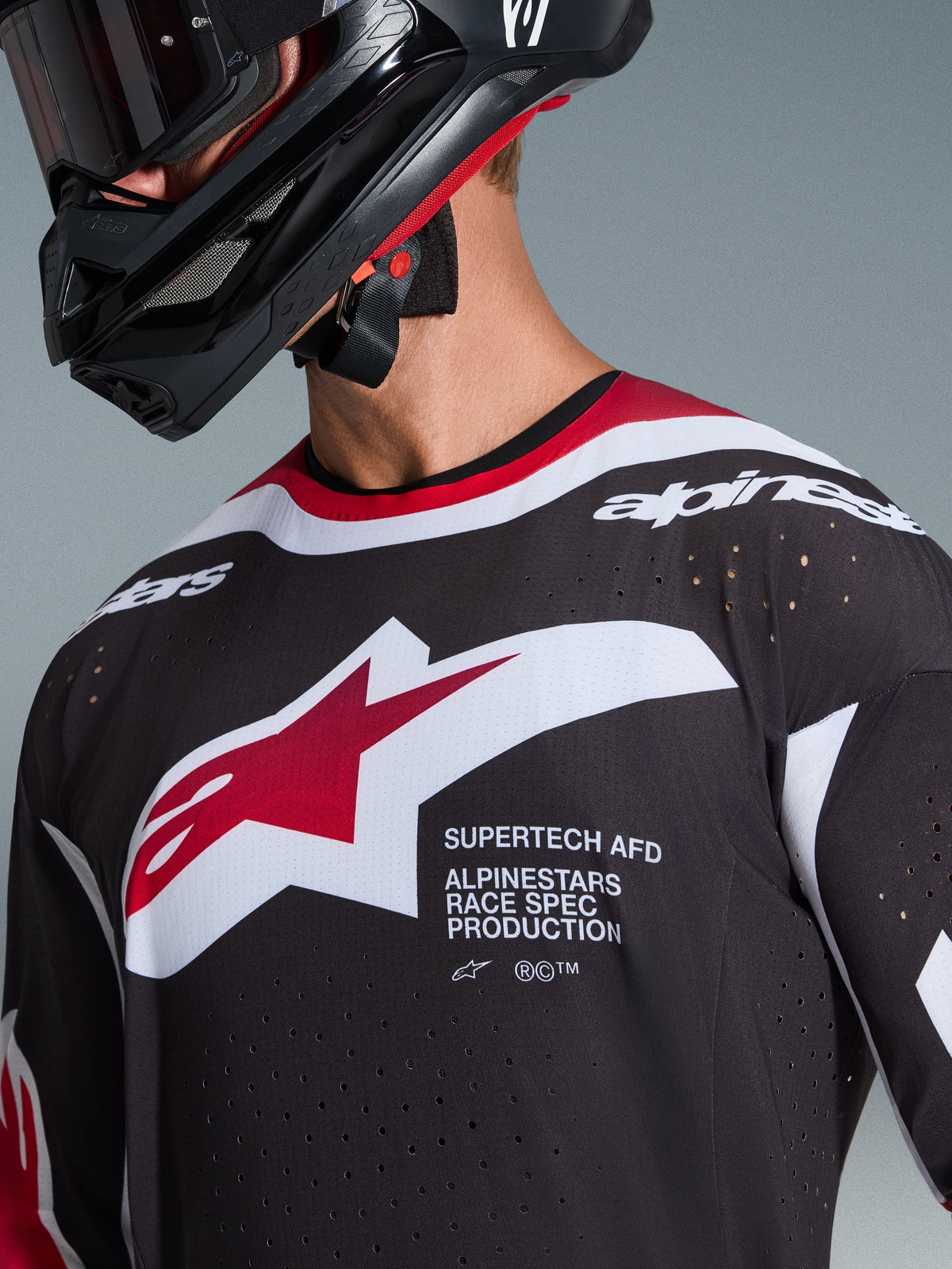 Alpinestars 2026 Supertech AFD Vista Jersey, primeiro plano de um piloto de motocross a usar uma camisola preta com detalhes em vermelho e branco, com um logótipo Alpinestars grande em branco e vermelho, perfurações de ventilação cortadas a laser e ajuste de competição profissional, combinada com um capacete preto.