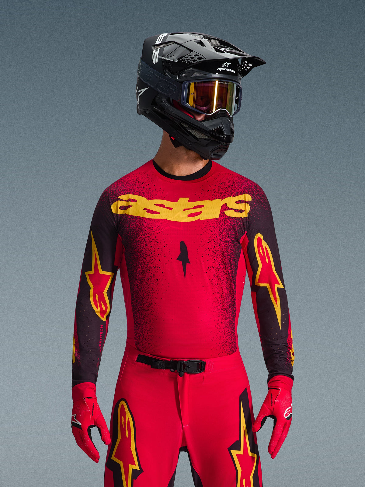 Conjunto de Equipamento Alpinestars 2026 Supertech Scenz, piloto de motocross com camisola vermelha, marca 'astars' amarela e detalhes pretos salpicados, calças vermelhas com logótipos de estrela amarelos nas coxas, luvas de corrida vermelhas e capacete preto com óculos amarelos refletores, equipamento de corrida profissional para performance em todo-o-terreno