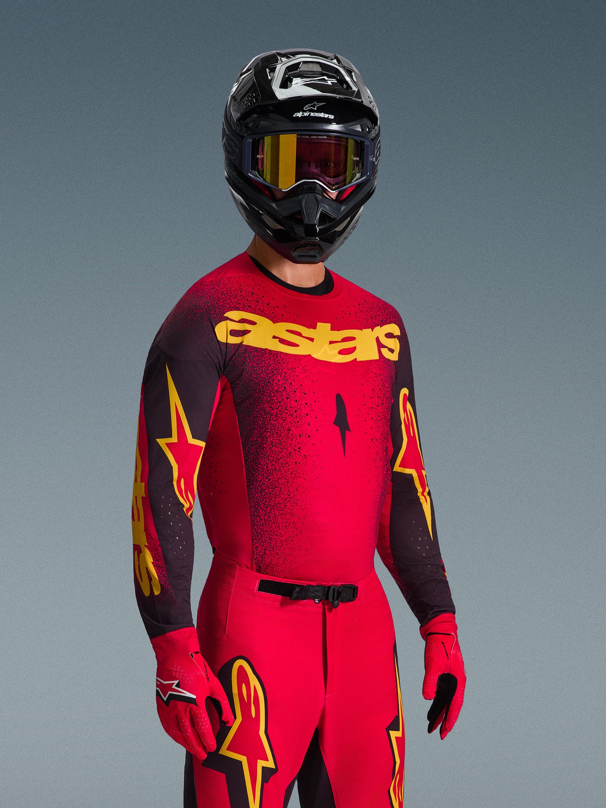 Piloto de motocross com o conjunto de equipamento Alpinestars Supertech Scenz 2026, camisola vermelha e preta com logótipo 'astars' amarelo e gradiente ponteado, calças vermelhas a condizer, luvas vermelhas e capacete preto com óculos amarelos, equipamento de corrida profissional para motocross
