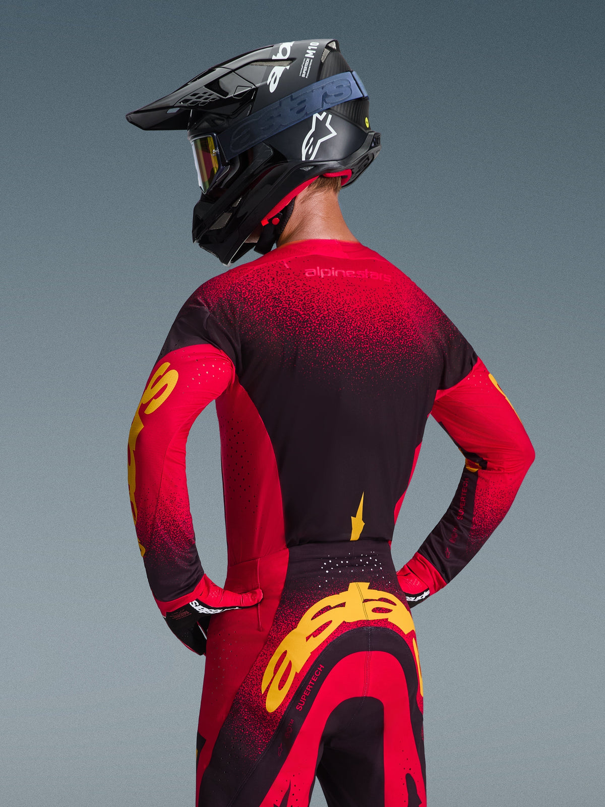 Camisola Alpinestars 2026 Supertech Scenz, vermelha, preta e amarela, vista traseira de piloto de motocross com equipamento de corrida a condizer, incluindo calças e capacete preto, design em gradiente vermelho e preto com logótipo Alpinestars em amarelo