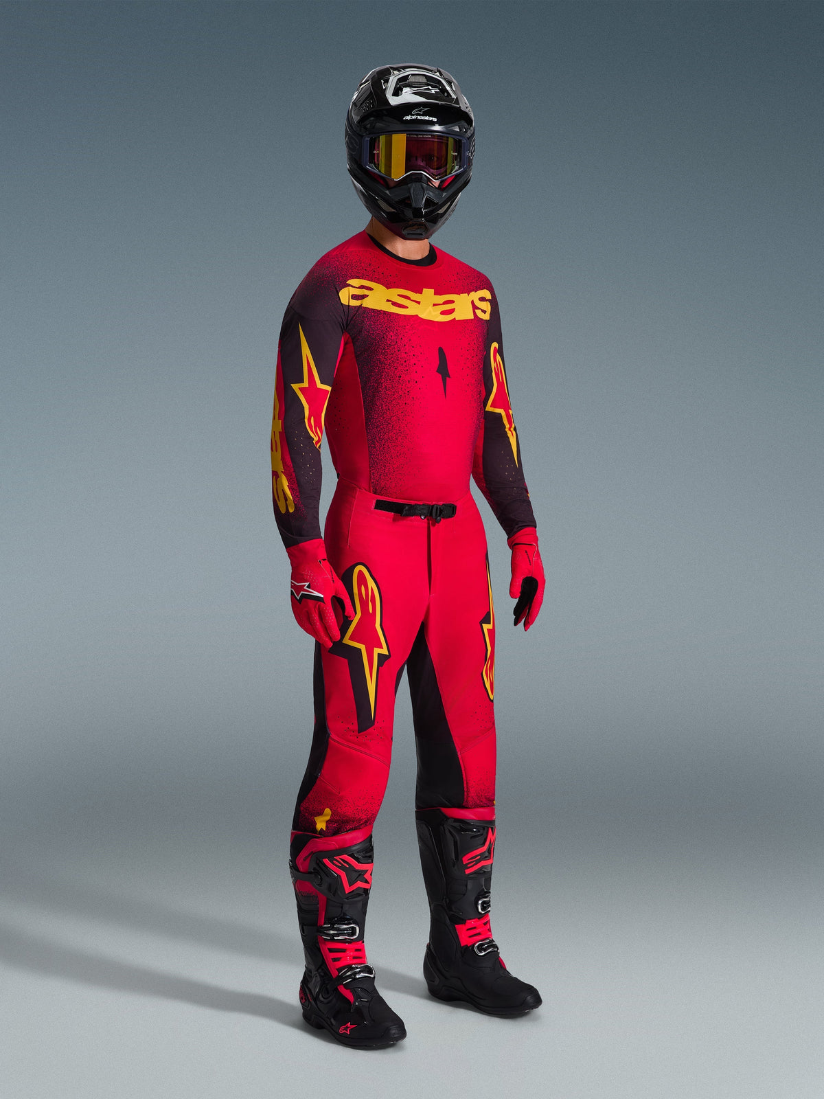 Piloto de motocross com a camisola Alpinestars Supertech Scenz 2026 em vermelho, preto e amarelo, calças vermelhas a condizer com detalhes em preto e amarelo, capacete preto com óculos, luvas vermelhas e botas pretas com detalhes vermelhos, conjunto completo de equipamento de corrida profissional