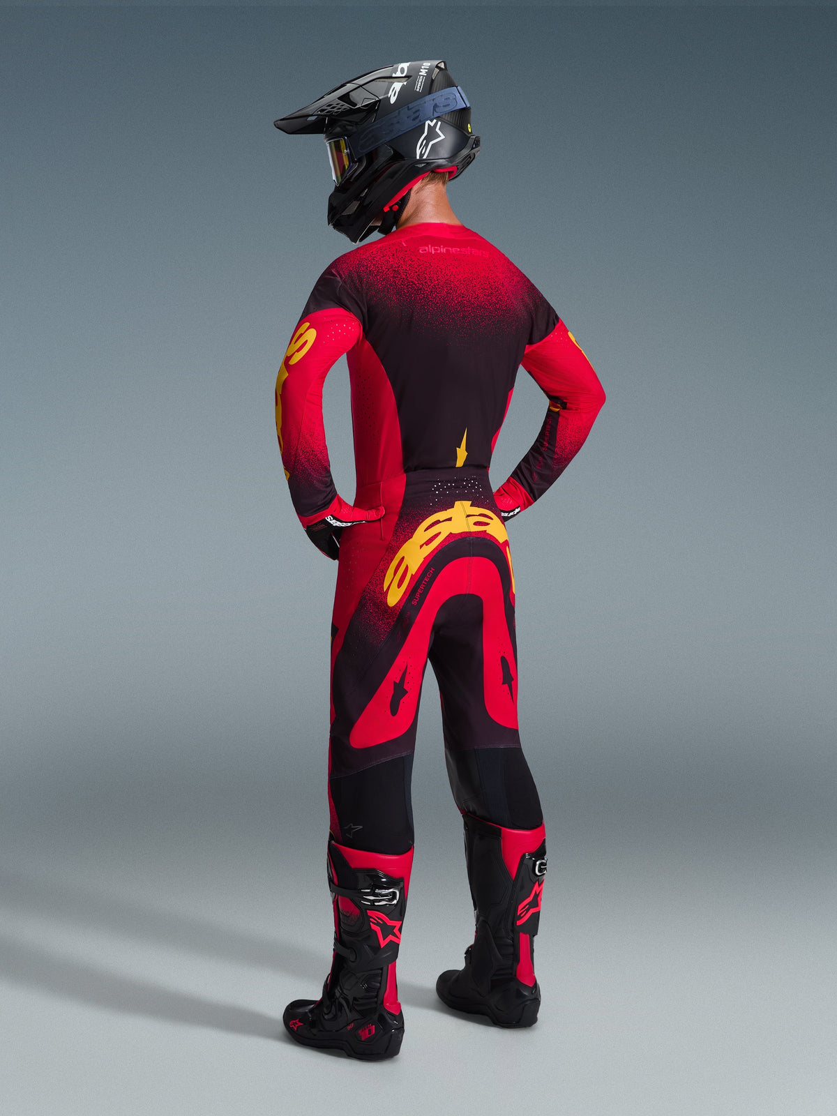 Piloto de motocross com equipamento de corrida completo visto de costas, camisola Alpinestars 2026 Supertech Scenz em vermelho, preto e amarelo, calças de motocross a condizer com logótipo amarelo, capacete preto e botas pretas e vermelhas, conjunto coordenado para competição profissional de todo-o-terreno