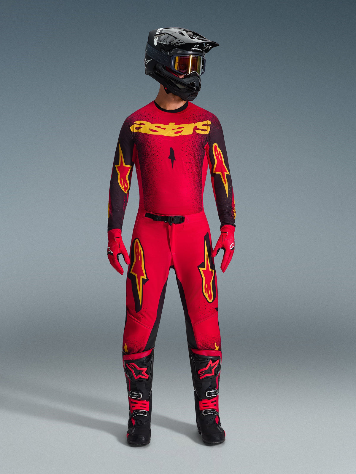 Piloto de motocross a usar a camisola Alpinestars 2026 Supertech Scenz em vermelho, preto e amarelo, combinada com calças a condizer, capacete preto com óculos, luvas vermelhas e botas pretas e vermelhas, conjunto de equipamento de competição profissional para performance todo-o-terreno