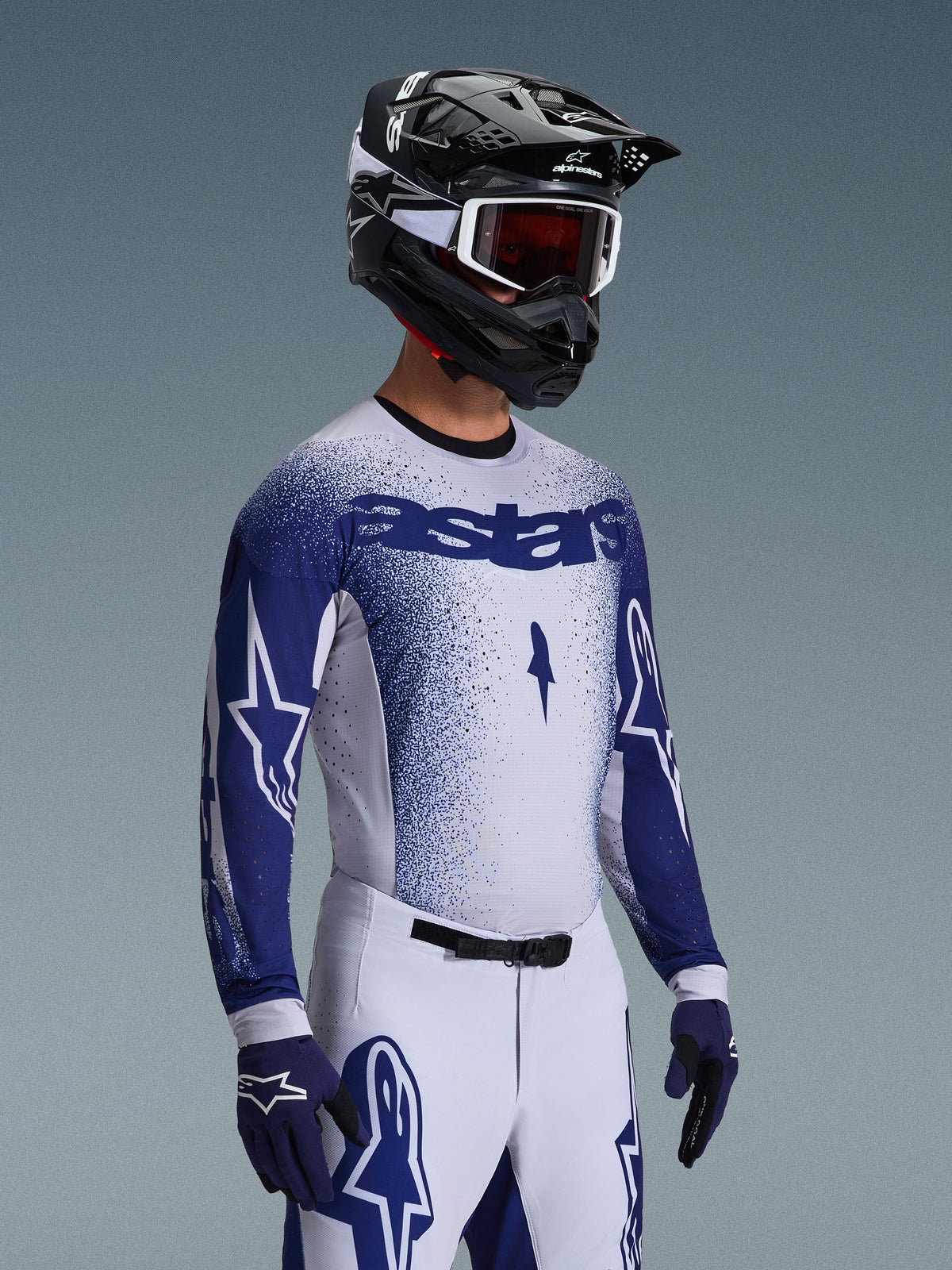 Piloto de motocross a usar a Camisola 2026 Supertech Scenz em cinzento claro e azul escuro, combinada com calças de corrida brancas e azuis a condizer, luvas azuis e um capacete preto com óculos brancos. A camisola apresenta um efeito de gradiente azul pontilhado no peito e nas mangas, corte atlético profissional e a marca 'astars' em destaque.