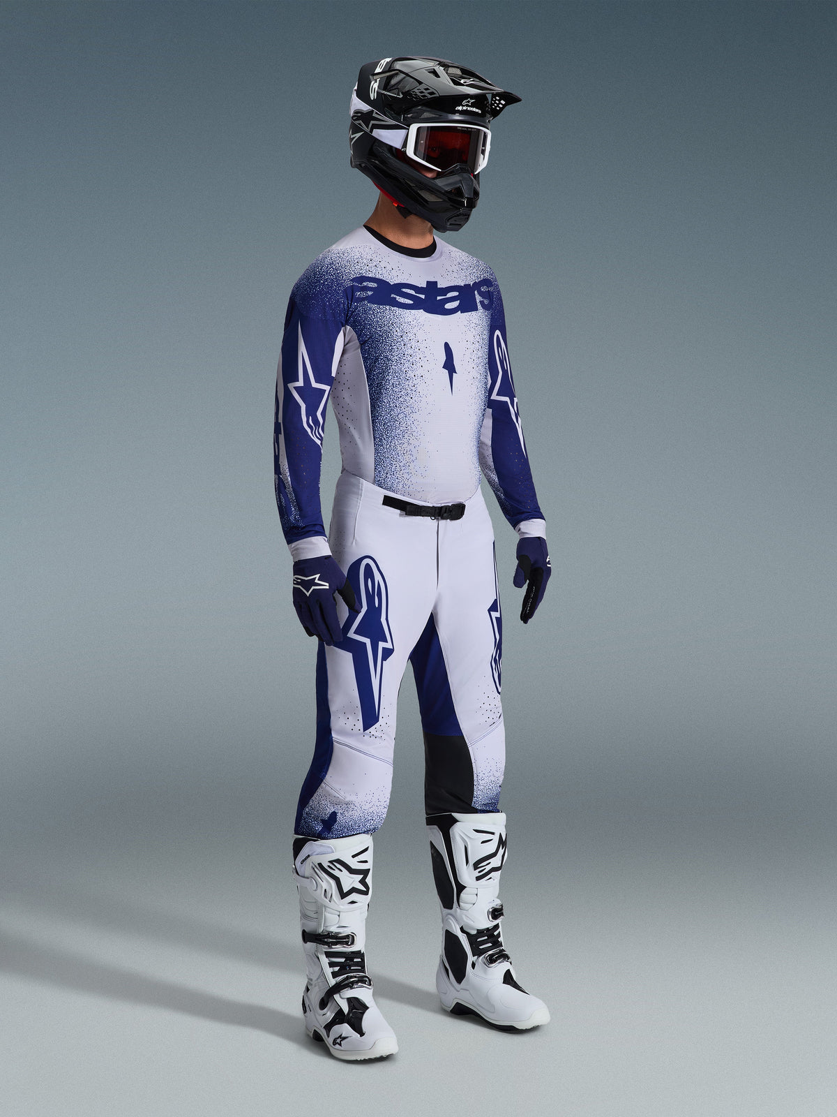 Piloto de motocross com equipamento profissional completo, camisola Supertech Scenz 2026 em cinzento claro e azul escuro, calças de motocross a combinar com grafismos de estrela, capacete preto com óculos vermelhos, botas de alta performance brancas e pretas, e luvas azuis e brancas coordenadas para competição off-road