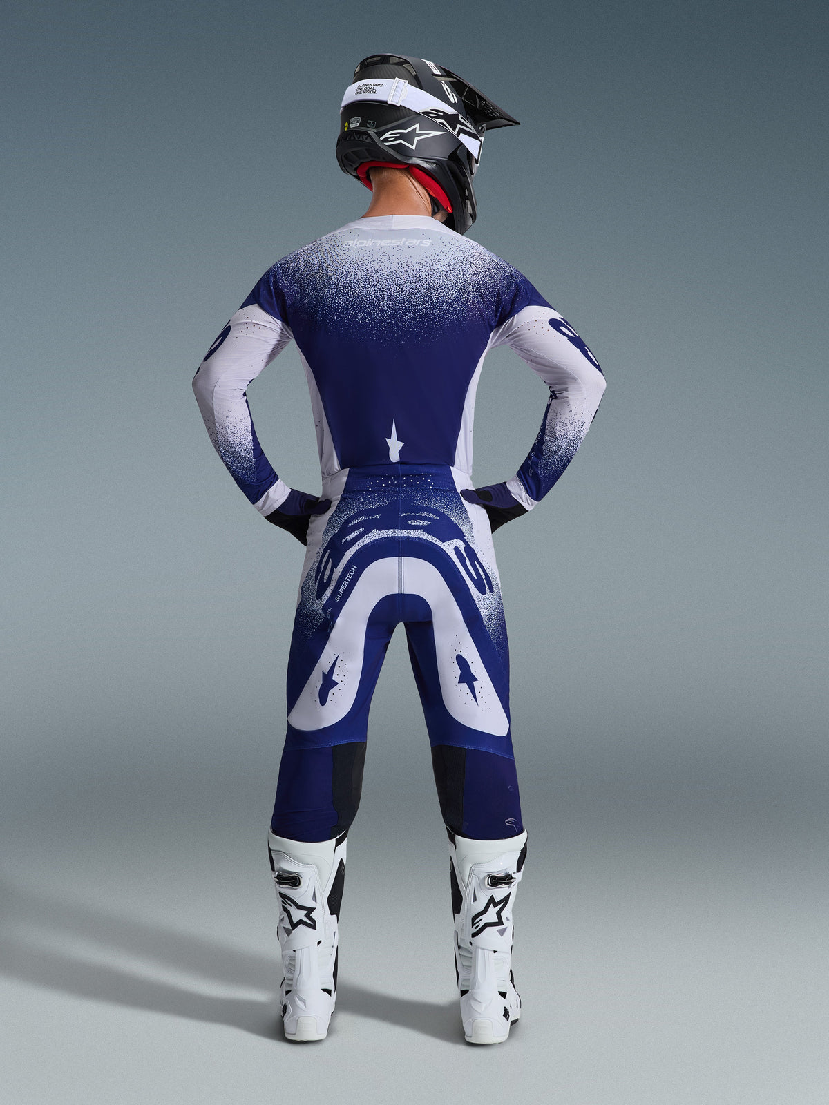 Piloto de motocross visto de trás com a camisola Alpinestars Supertech Scenz 2026, cinzento claro e azul escuro com um design de gradiente pontilhado, calças de motocross a condizer, botas de corrida brancas e capacete preto, equipamento completo de corrida todo-o-terreno.