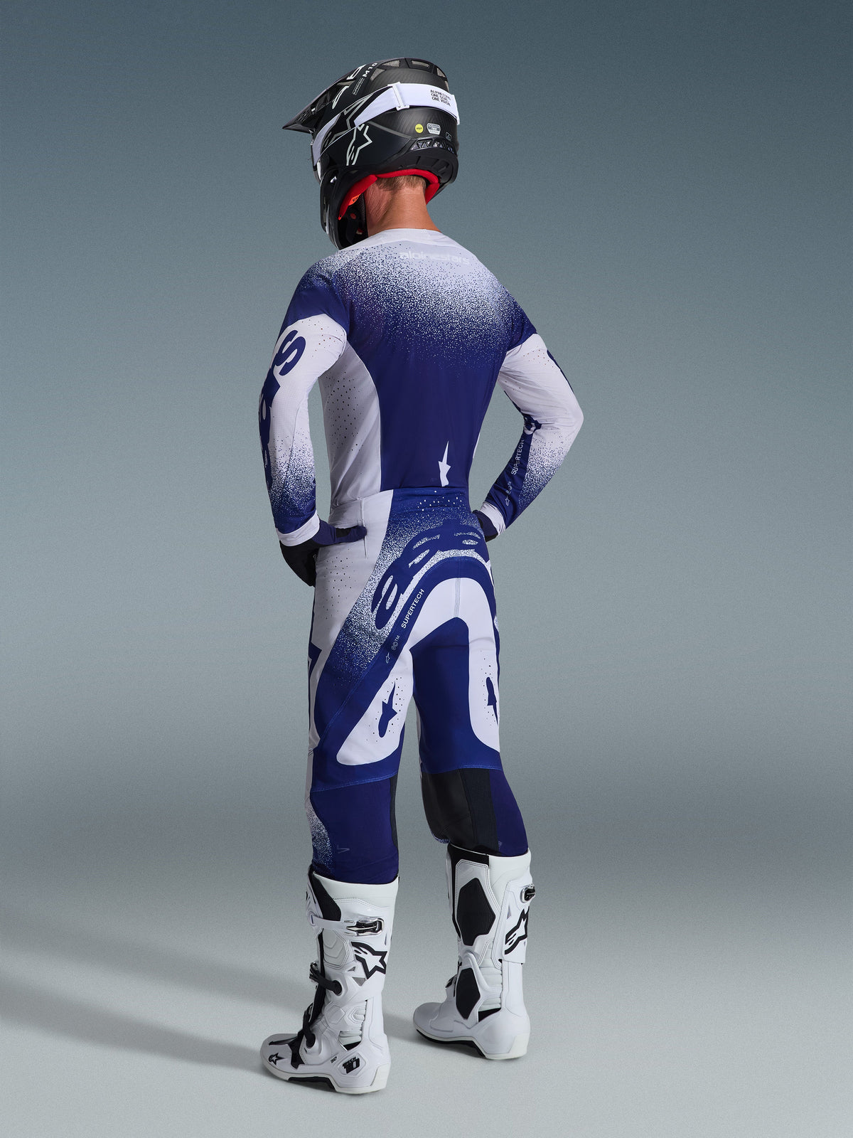 Motocross rider wearing the Alpinestars 2026 Supertech Scenz Gear Set, navy blue and white jersey and pants with a speckled gradient effect, rear view showing athletic fit, paired with white and black boots and a black helmet, professional racing gear for off-road performance