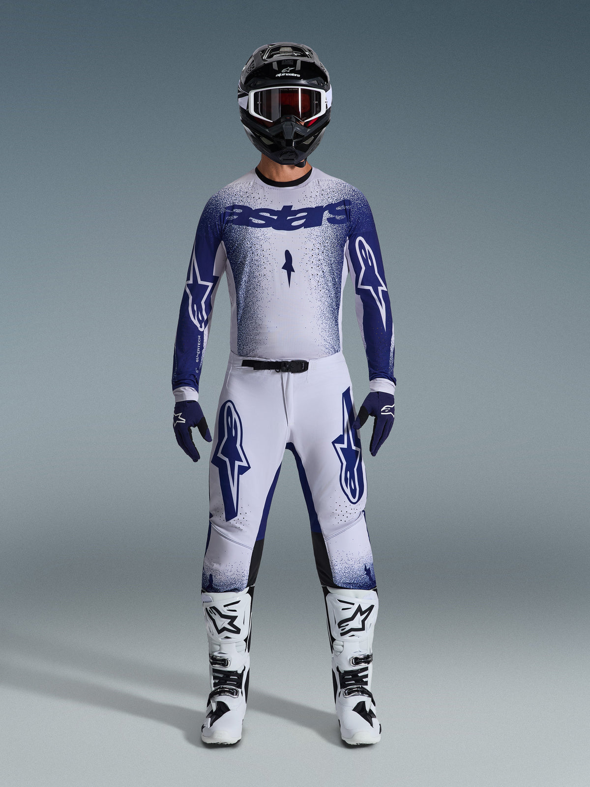Piloto de motocross com equipamento completo, camisola Supertech Scenz 2026 em cinzento claro e azul escuro, calças cinzentas claras a condizer com logótipos azuis, capacete preto e branco com óculos vermelhos, botas brancas e pretas, luvas azuis escuras, conjunto coordenado para corridas de todo-o-terreno