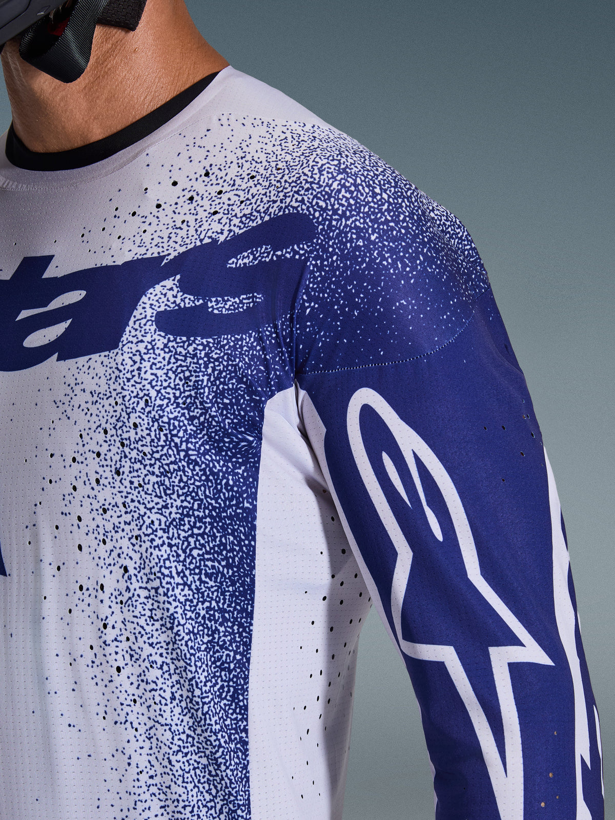 Alpinestars 2026 Supertech Scenz Jersey, Light Gray and Dark Blue, close-up of shoulder and chest showing laser-cut perforations, white Astars logo on blue sleeve, speckled graphic design, professional motocross racing gear.