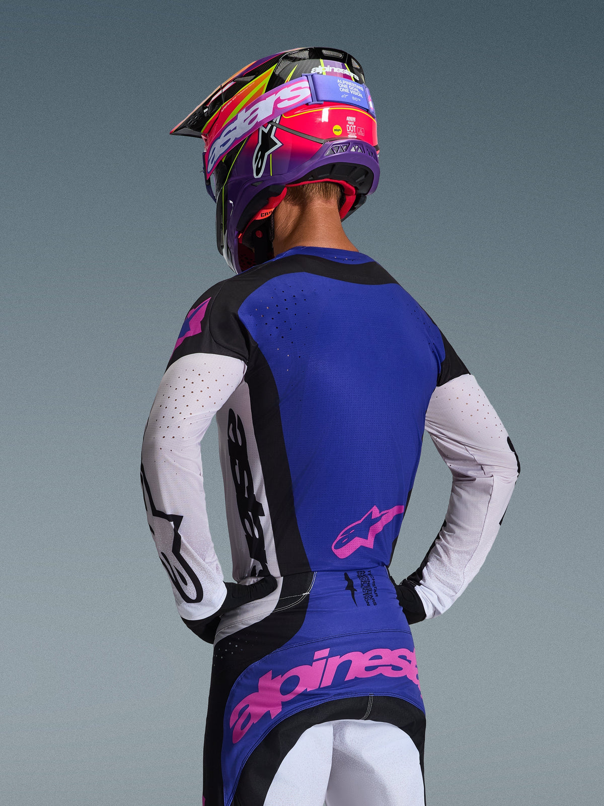 Piloto de motocross visto de costas com a Camisola Techstar Knif 2026, em preto, cinzento claro e roxo, com mangas perfuradas brancas e logótipos Alpinestars cor-de-rosa, combinada com um capacete roxo e rosa condizente, equipamento profissional de competição todo-o-terreno.