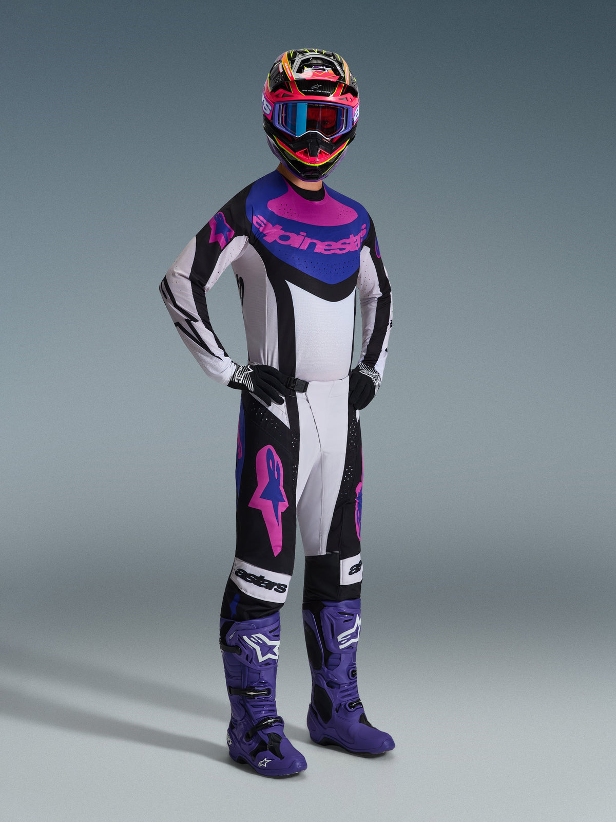 Piloto de motocross com a camisola Alpinestars 2026 Techstar Knif em preto, cinzento claro e roxo, calças a condizer com logótipos cor-de-rosa grandes, botas roxas e capacete multicolorido com óculos azuis, equipamento de corrida completo para performance em todo-o-terreno