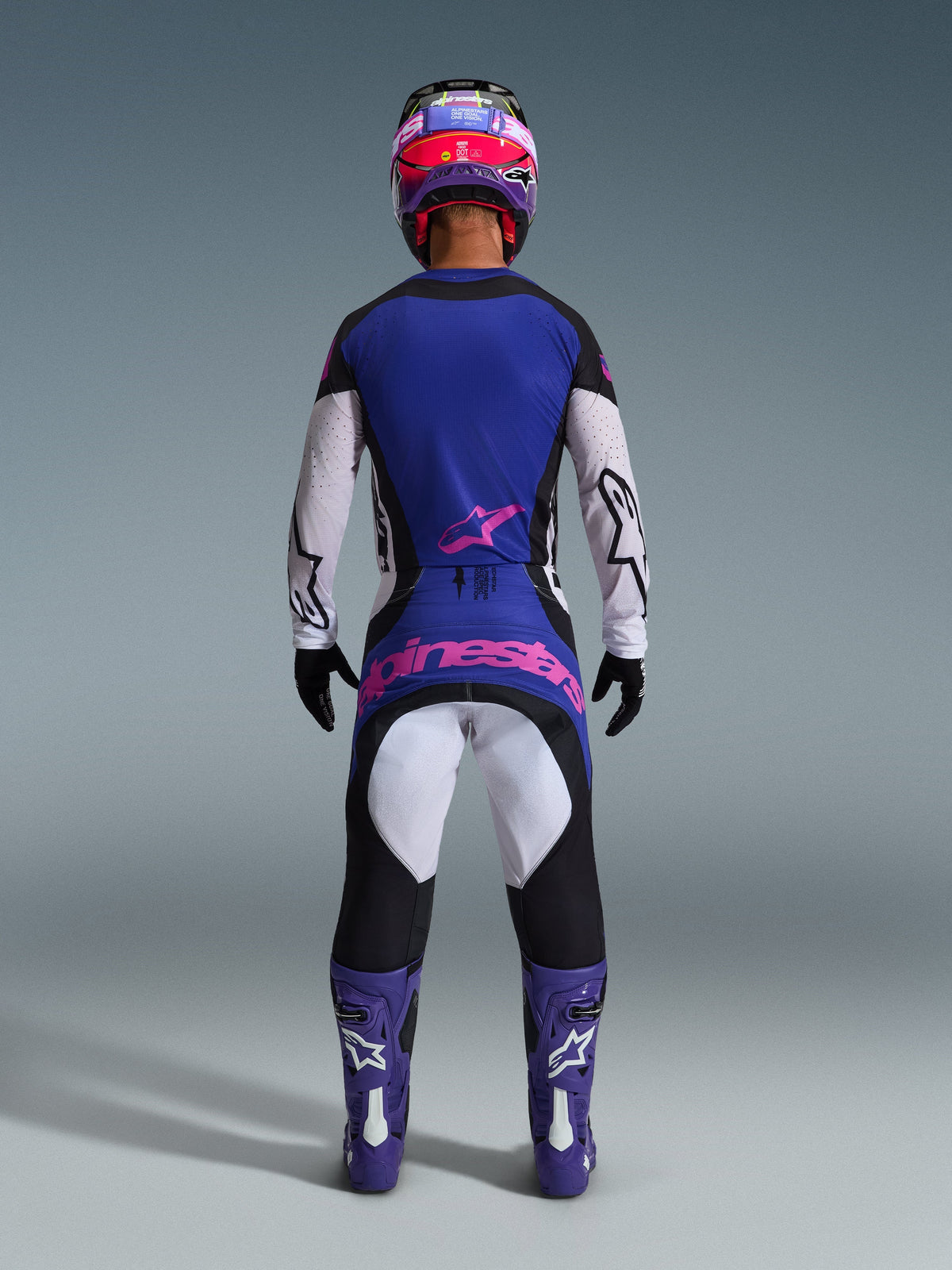 Vista traseira de um piloto de motocross a usar a Camisola Alpinestars 2026 Techstar Knif em preto, cinzento claro e roxo, com mangas brancas e um logótipo rosa na parte inferior das costas, combinada com calças de motocross a condizer, botas roxas e um capacete colorido num cenário de estúdio.