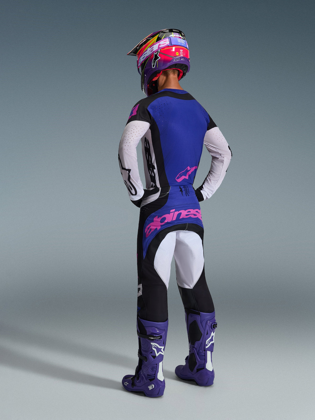 Piloto de motocross visto de costas com equipamento completo Alpinestars, incluindo a camisola Techstar Knif 2026 com costas azuis e mangas brancas perfuradas, calças pretas com logótipo em cor-de-rosa, capacete colorido e botas roxas, conjunto coordenado para competição de todo-o-terreno