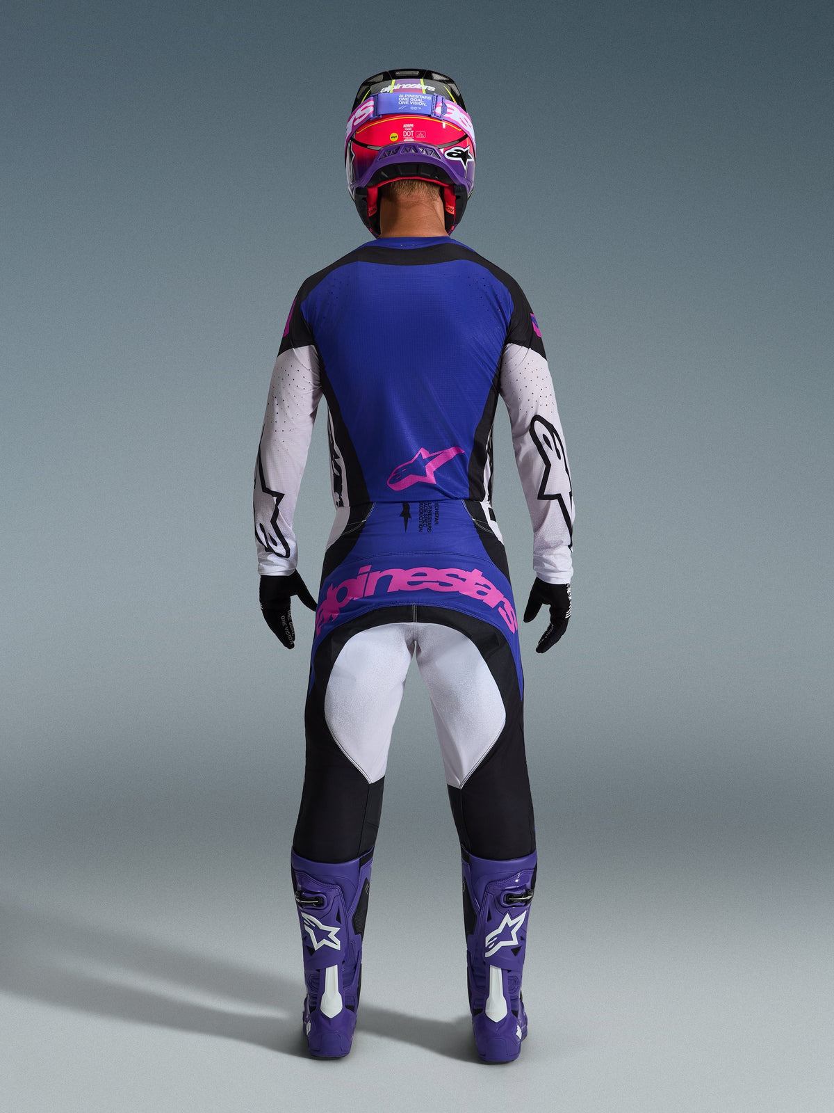 Piloto de motocross visto de costas a usar a Camisola Techstar Knif 2026, em preto, cinzento claro e roxo, calças de motocross a combinar com logótipo cor-de-rosa, botas e capacete roxos, equipamento de todo-o-terreno de alta performance coordenado
