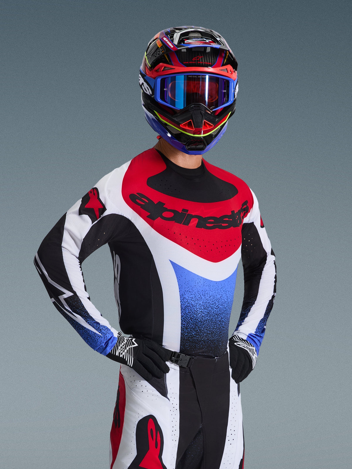 Piloto de motocross a usar a camisola Alpinestars 2026 Techstar Knif em vermelho, branco e azul, com logótipo preto no peito, combinada com calças de motocross a condizer, capacete de corrida coordenado com óculos azuis e luvas pretas e brancas, equipamento profissional de todo-o-terreno para competição