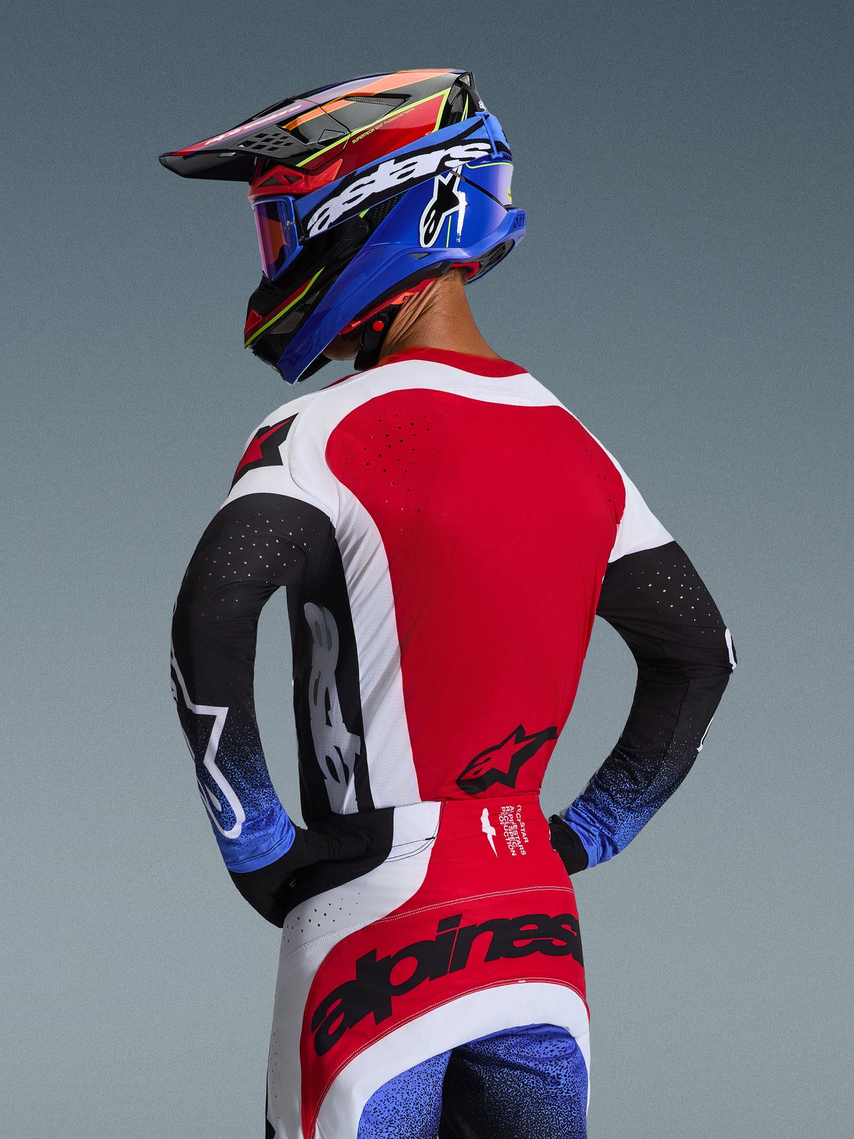 Piloto de motocross visto de costas a usar a Camisola Techstar Knif 2026 em vermelho, branco e preto, com perfurações a laser nas costas e logótipos em destaque, combinada com calças a condizer com detalhes azuis e capacete Alpinestars azul e preto, estética de corrida profissional em todo-o-terreno