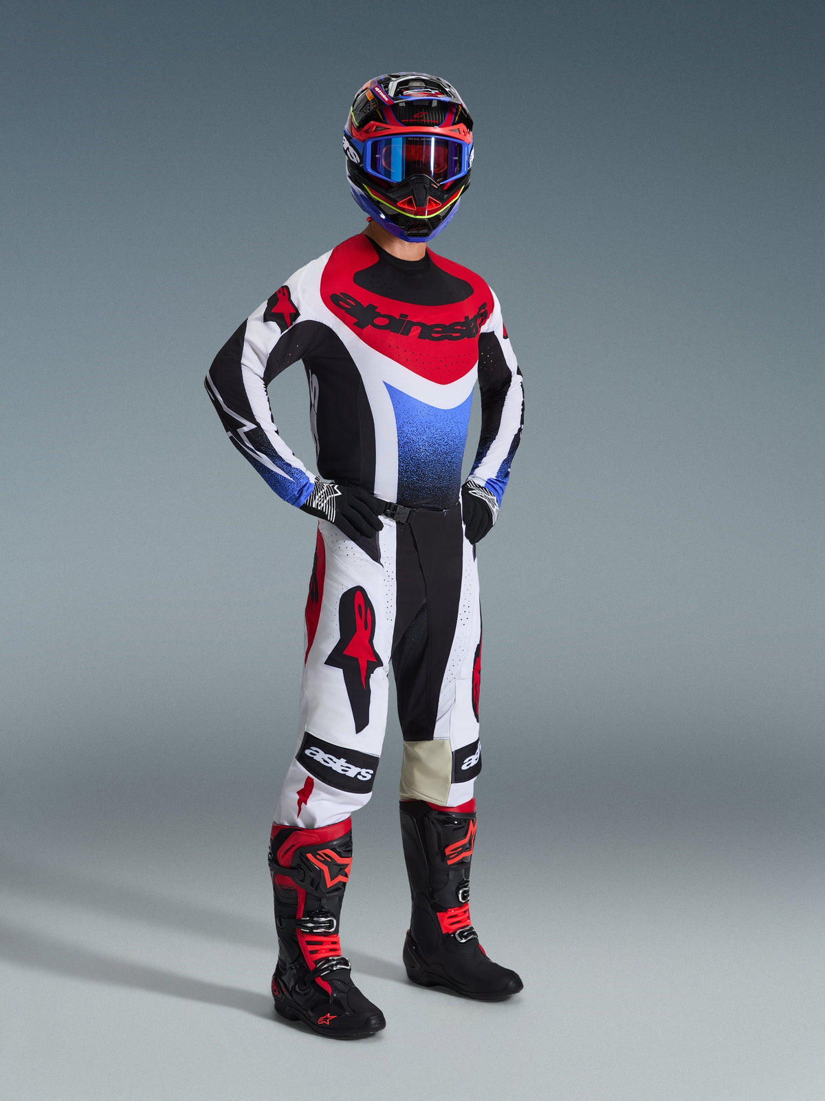 Motocross rider wearing the Alpinestars 2026 Techstar Knif Gear Set, jersey featuring black white and red panels with a blue gradient chest, white pants with red Alpinestars logos and black accents, black and red boots, complete professional off-road racing ensemble.