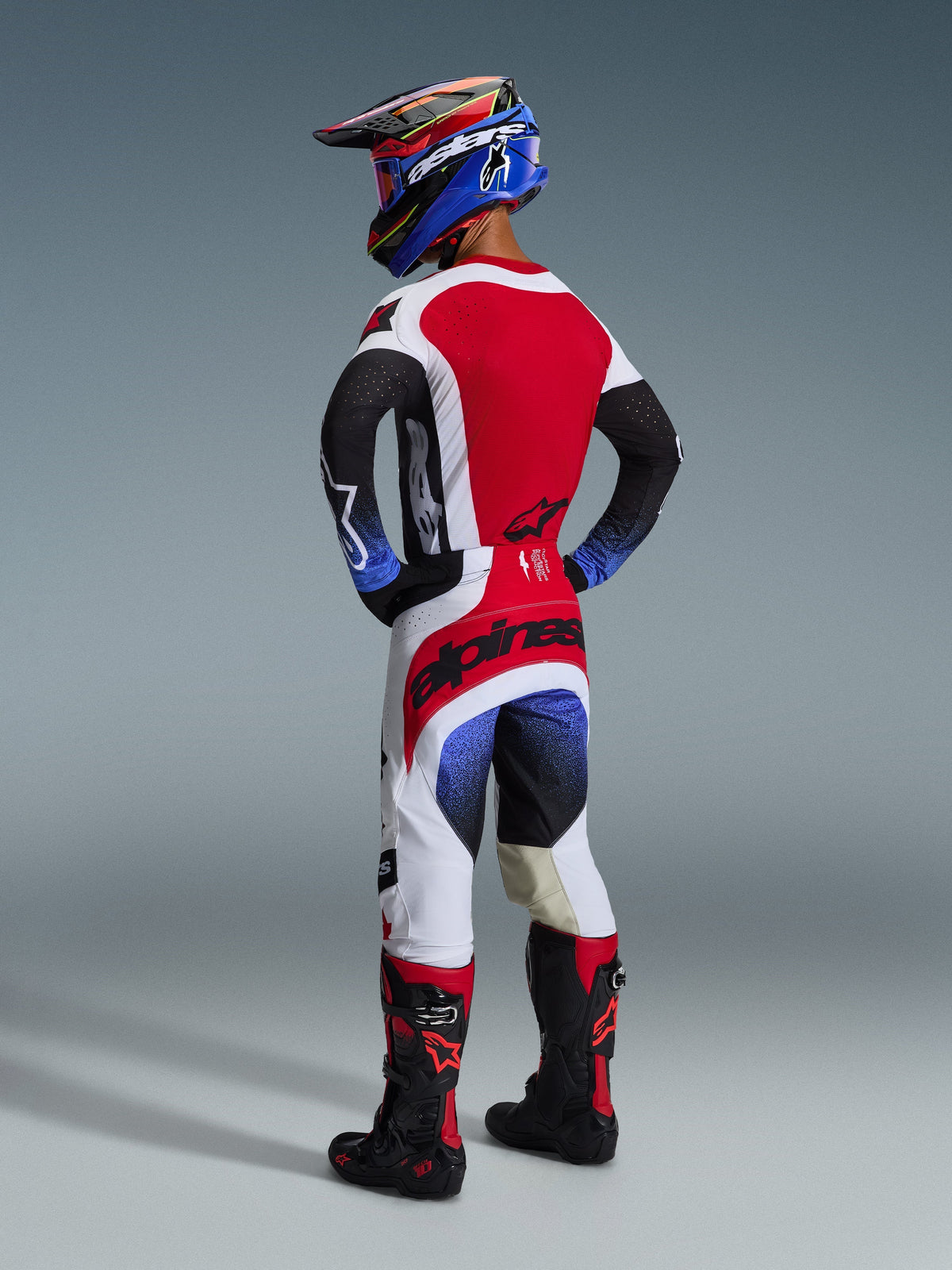 Motocross rider from a rear view wearing the 2026 Techstar Knif Gear Set, Black White and Red jersey with blue gradient sleeves and laser-cut perforations, matching white and red pants with bold black Alpinestars branding on the seat, blue and black helmet with red goggles, black and red motocross boots, complete off-road racing kit.
