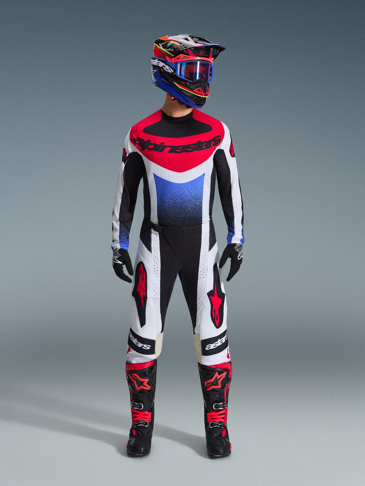Alpinestars 2026 Techstar Knif Jersey, camisola de motocross em branco, preto, vermelho e azul com um gráfico em gradiente azul, usada como parte de um kit completo de corrida todo-o-terreno, incluindo calças a condizer, botas pretas e vermelhas e um capacete coordenado com óculos.