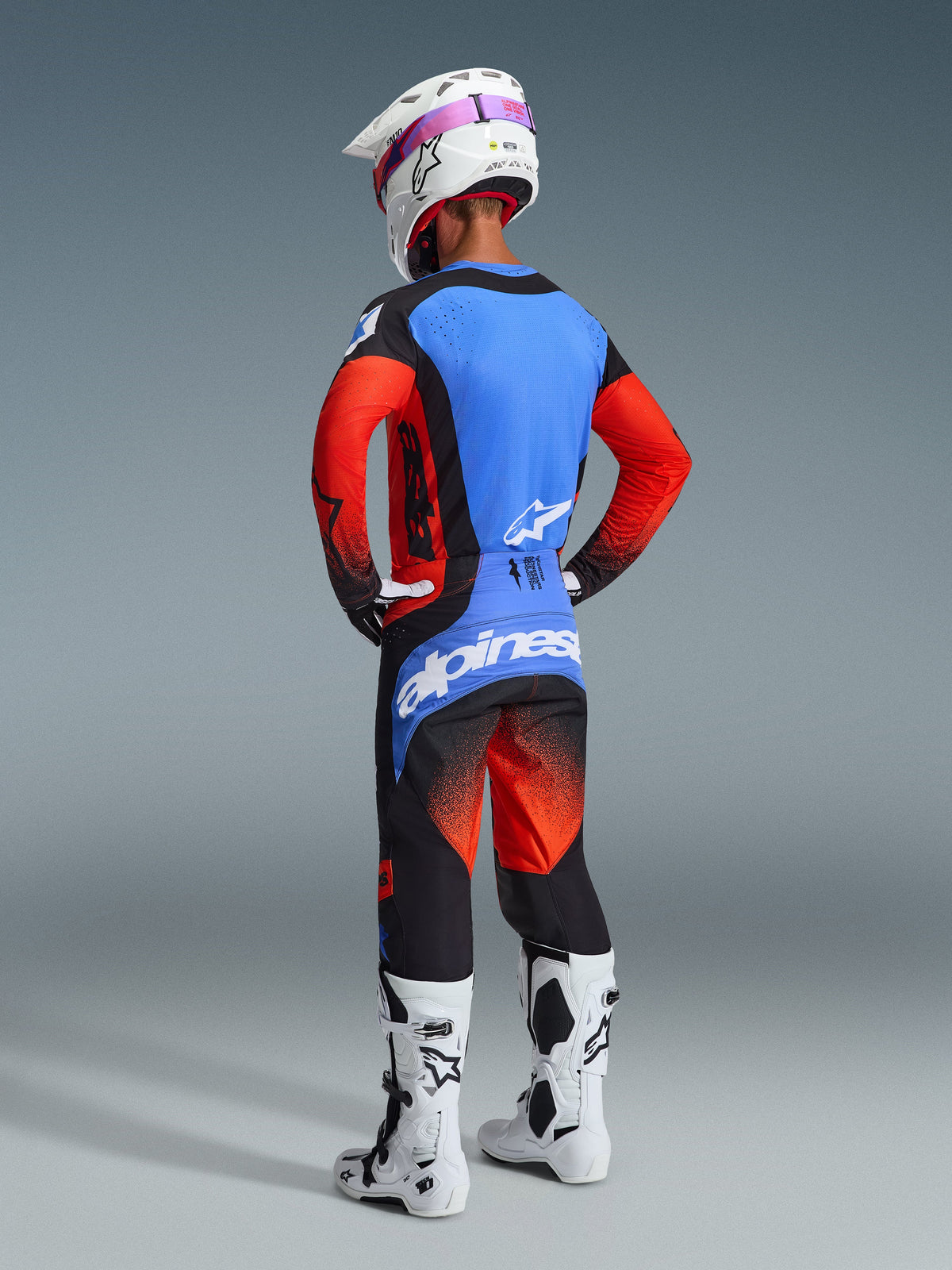 Vista traseira de um piloto de motocross com o Conjunto de Equipamento Alpinestars 2026 Techstar Knif, composto por uma camisola azul e vermelha com detalhes pretos, calças pretas e vermelhas com um grande logótipo azul no assento, capacete branco e botas de motocross brancas, kit profissional de corrida de todo-o-terreno.