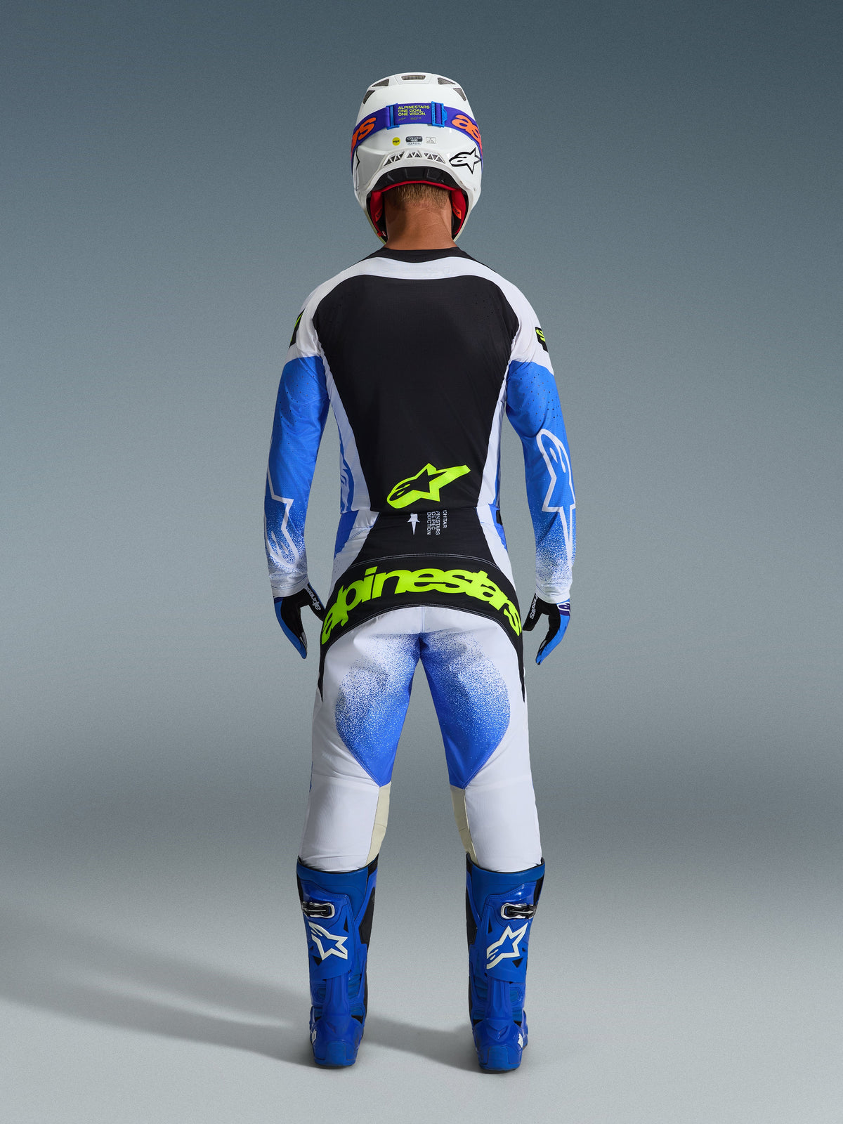 Conjunto de Equipamento Alpinestars 2026 Techstar Knif, vista traseira de um piloto de motocross em Azul Ucla, Preto e Branco, com logótipos em Amarelo Fluo na camisola e nas calças, combinado com botas de corrida azuis e capacete branco.