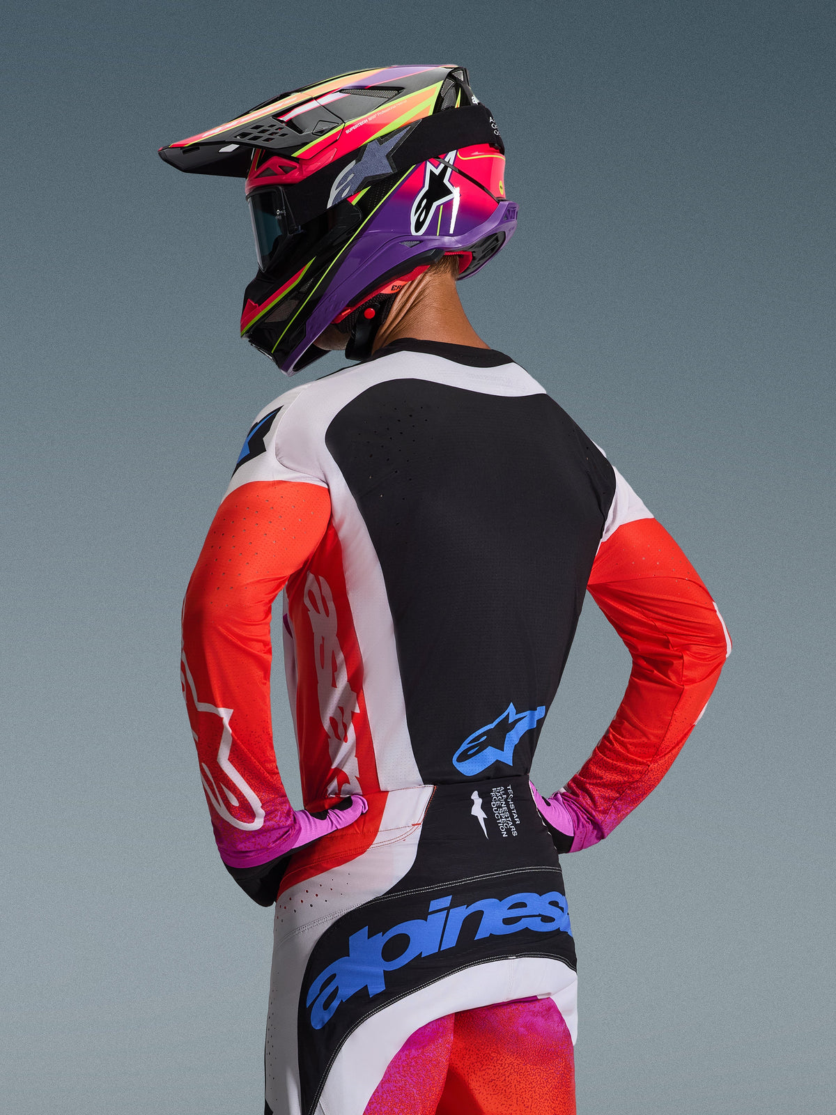 Motocross rider from behind wearing the Alpinestars 2026 Techstar Knif Jersey in Light Gray Orange Fluo Black, featuring a black back panel with laser-cut ventilation, orange sleeves, and light gray side panels, coordinated with matching pants and a colorful helmet.