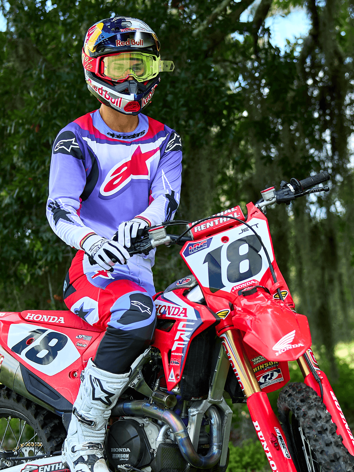 Piloto de motocross numa moto Honda CRF vermelha, a usar a camisola Alpinestars 2025 Fluid Wurx em roxo e vermelho brilhante, calças a condizer em roxo e vermelho, capacete Red Bull com óculos amarelos, botas e luvas Alpinestars brancas, equipamento profissional de MX para condução todo-o-terreno