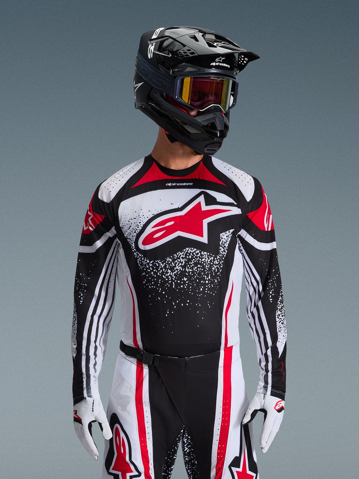 Alpinestars 2026 Techstar Nomur Jersey, camisola de motocross, preta, branca e vermelha, usada por um piloto com calças a condizer, capacete preto e luvas brancas, com um grande logótipo Astars vermelho e design gráfico salpicado, corte atlético de corrida para competição todo-o-terreno