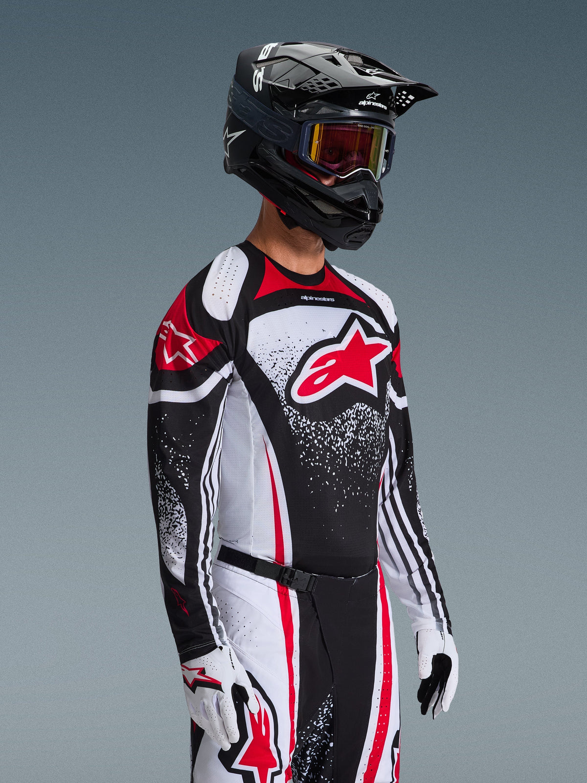 Piloto de motocross a usar a camisola Alpinestars 2026 Techstar Nomur em preto, branco e vermelho, com um grande logótipo Astars vermelho e padrões gráficos salpicados, combinada com calças de corrida a condizer, luvas brancas e capacete preto com óculos, conjunto coordenado para competição todo-o-terreno