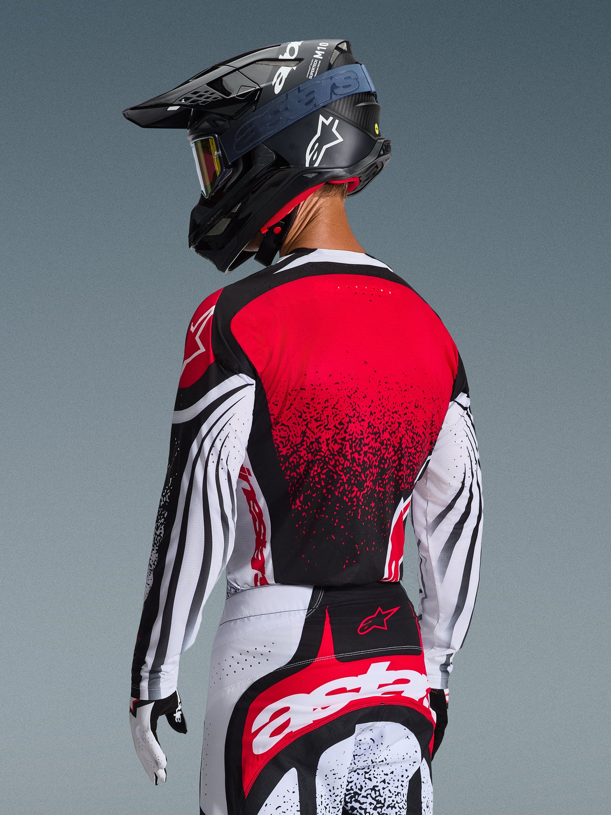 Piloto de motocross visto de costas a usar a Camisola Techstar Nomur 2026, com costas vermelhas e design em gradiente preto, mangas brancas com detalhes em preto, calças vermelhas e brancas a condizer, capacete preto e luvas brancas, equipamento profissional de corrida todo-o-terreno.