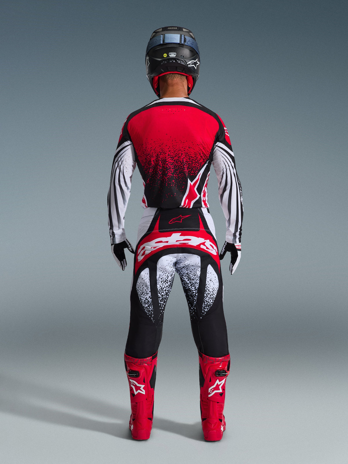 Piloto de motocross visto de costas a usar a camisola Alpinestars Techstar Nomur 2026, vermelha, preta e branca com padrão ponteado, calças de corrida a condizer, botas de motocross vermelhas e capacete preto, conjunto completo de equipamento para competição de todo-o-terreno