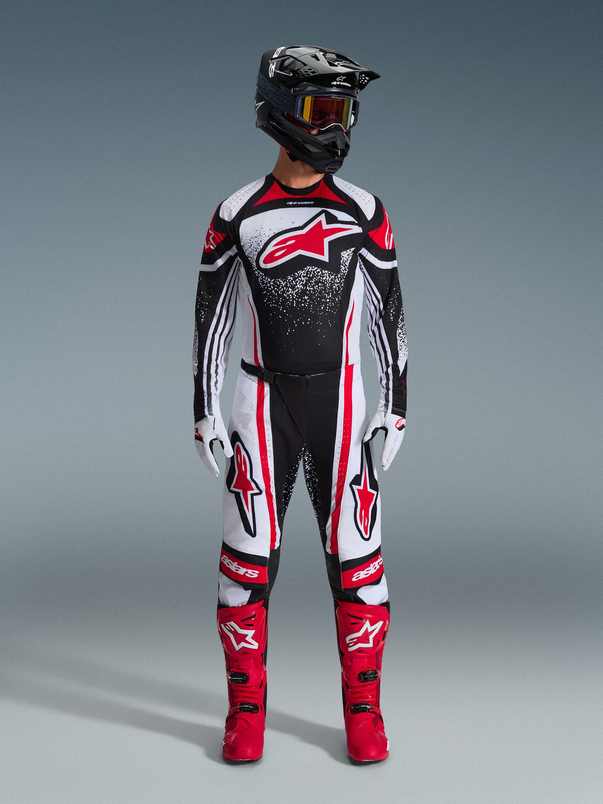 Piloto de motocross com equipamento completo de corrida Alpinestars, Camisola Techstar Nomur 2026 em Preto Branco Vermelho, calças a condizer com detalhes vermelhos, capacete preto e botas de cor vermelha vibrante, apresentando um grande logótipo Astars vermelho no peito com padrão de gradiente salpicado, conjunto coordenado para competição de todo-o-terreno