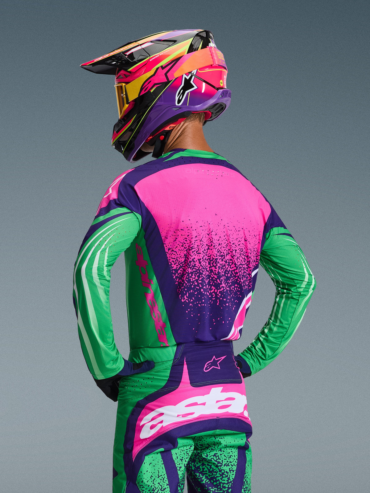 Piloto de motocross visto de costas com o Conjunto de Equipamento Alpinestars 2026 Techstar Nomur, camisola e calças em Deep Purple Green e Pink Fluo, com gráficos arrojados e corte atlético de corrida, incluindo capacete multicolorido e óculos amarelos.
