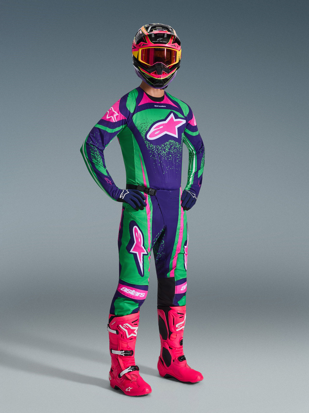Piloto de motocross com o conjunto Alpinestars 2026 Techstar Nomur em roxo escuro, verde e rosa fluo, com camisola e calças a condizer, capacete colorido com óculos cor-de-rosa e botas cor-de-rosa vibrantes, equipamento profissional de competição todo-o-terreno