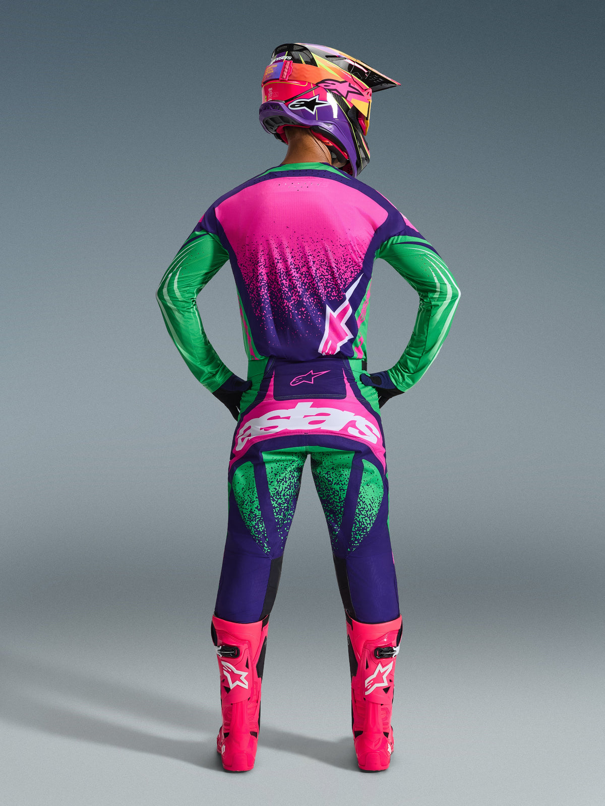 Alpinestars 2026 Techstar Nomur Gear Set, piloto de motocross visto de costas com camisola e calças em roxo escuro, verde e rosa fluo, capacete multicolorido a condizer, botas rosa fluo vibrante, equipamento de corrida profissional coordenado para performance todo-o-terreno