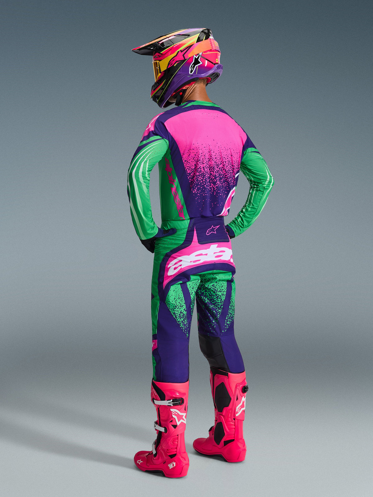 Piloto de motocross a usar o conjunto de equipamento Alpinestars Techstar Nomur 2026, nas cores roxo escuro, verde e rosa fluorescente, camisola com costas cor-de-rosa e textura pontilhada, calças a condizer com logótipos brancos, botas rosa néon vibrantes e capacete colorido, vista traseira de corpo inteiro para performance em corridas todo-o-terreno