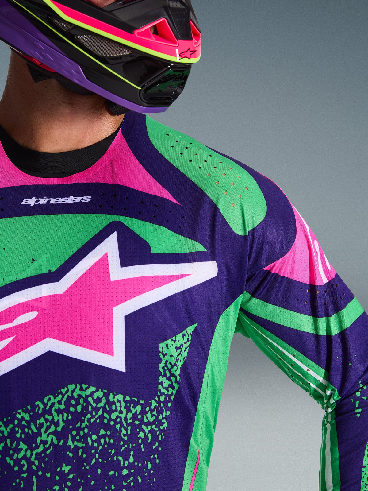 Primeiro plano da camisola Alpinestars Techstar Nomur 2026, em roxo profundo com detalhes em verde e rosa fluo, apresentando um grande logótipo de estrela cor-de-rosa no peito e perfurações cortadas a laser no ombro, vestida por um piloto com um capacete de motocross em roxo e rosa a condizer.