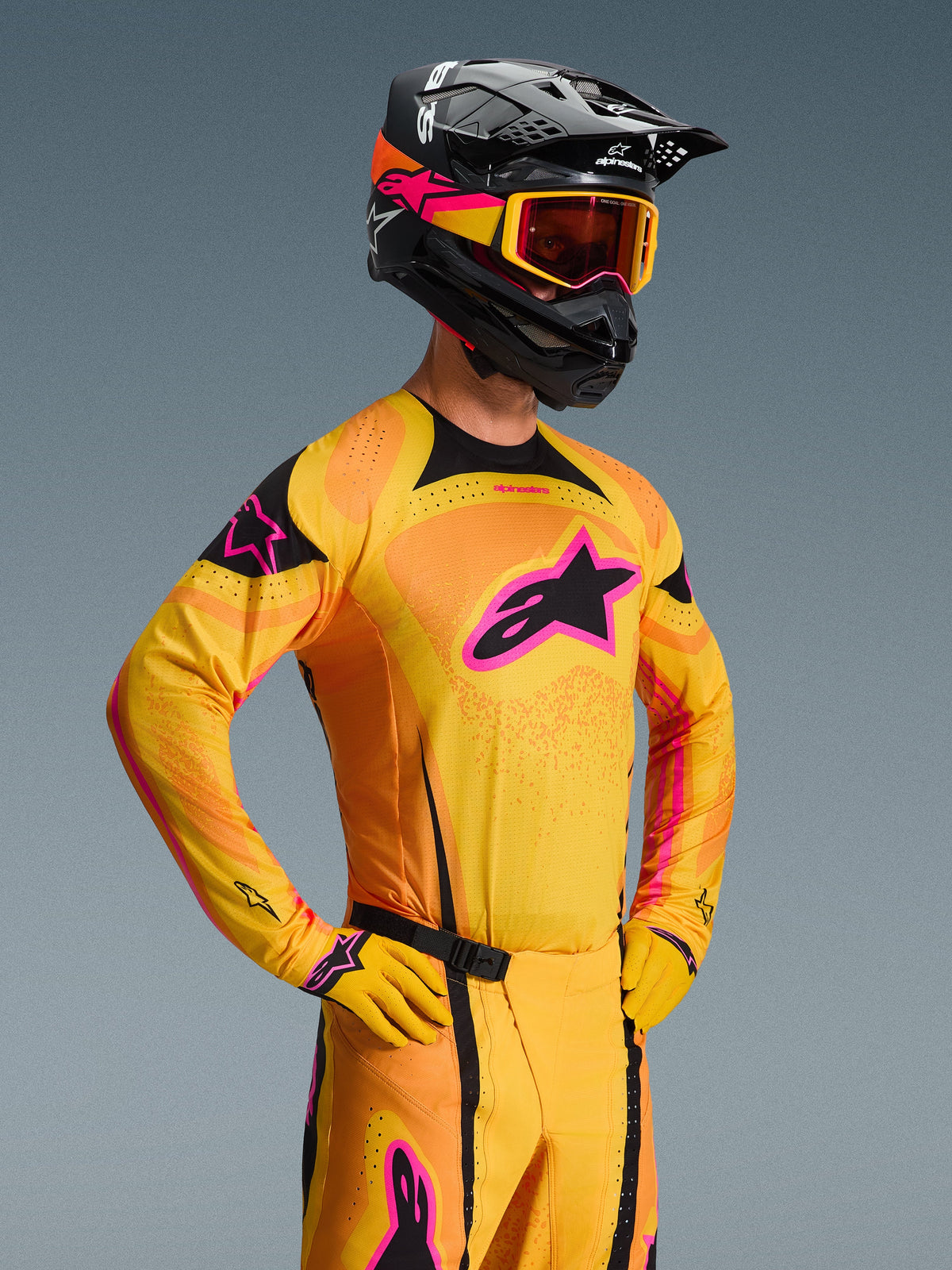 Alpinestars 2026 Techstar Nomur Jersey, camisola de motocross, laranja, amarela e rosa, usada por um piloto com capacete preto e óculos a condizer, com um grande logótipo de estrela cor-de-rosa no peito, detalhes pretos e detalhes de ventilação cortados a laser