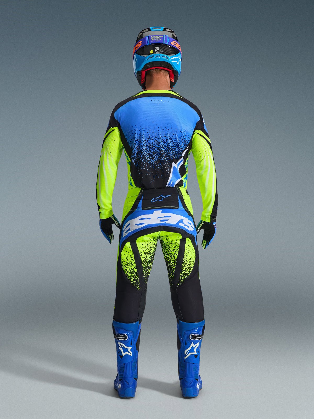 Vista traseira de um piloto de motocross a usar a Camisola Techstar Nomur 2026 em Azul Marinho Escuro, Amarelo Fluo e Azul UCLA, combinada com calças de motocross a condizer em amarelo e preto com a marca 'Astars', botas de corrida azuis e um capacete coordenado, equipamento profissional completo para corridas todo-o-terreno.