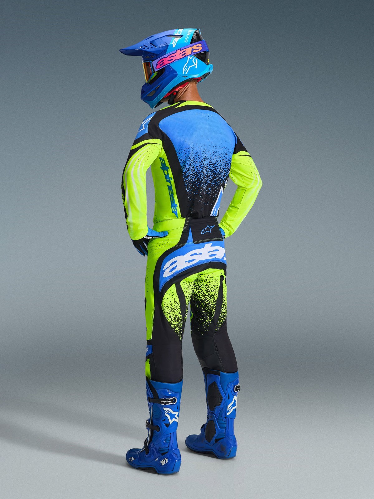 Piloto de motocross de costas a usar equipamento de corrida completo, Camisola Techstar Nomur 2026 em amarelo fluo e azul, calças amarelas e pretas com detalhes azuis, capacete azul com logótipo Alpinestars rosa e botas azuis, foto de estúdio realçando o design traseiro e o corte atlético.
