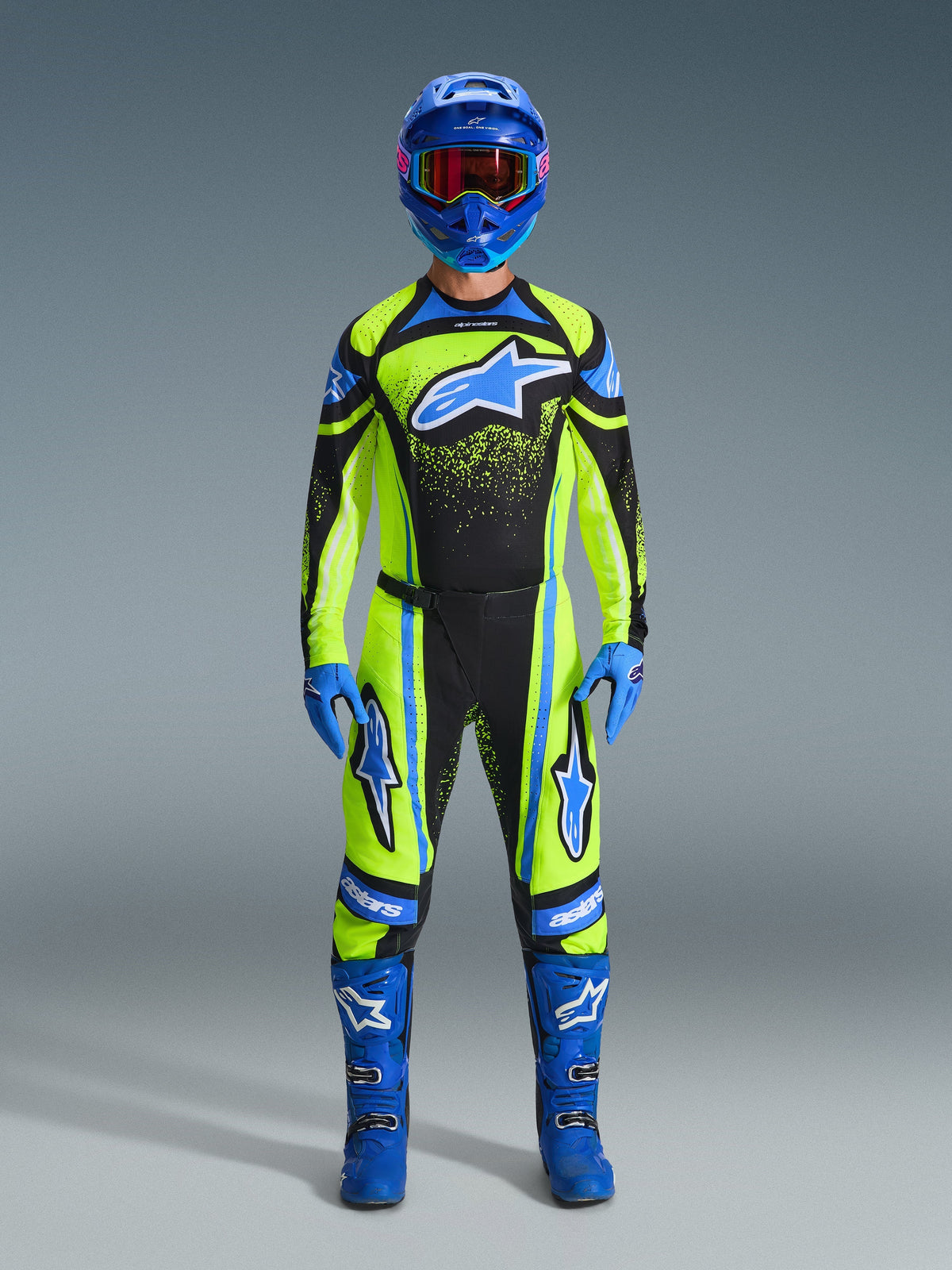 Piloto de motocross com equipamento completo Alpinestars, camisola Techstar Nomur 2026 em amarelo fluo e azul escuro, calças de motocross a condizer, capacete azul com óculos de lentes vermelhas, luvas e botas azuis, conjunto profissional para corridas de todo-o-terreno