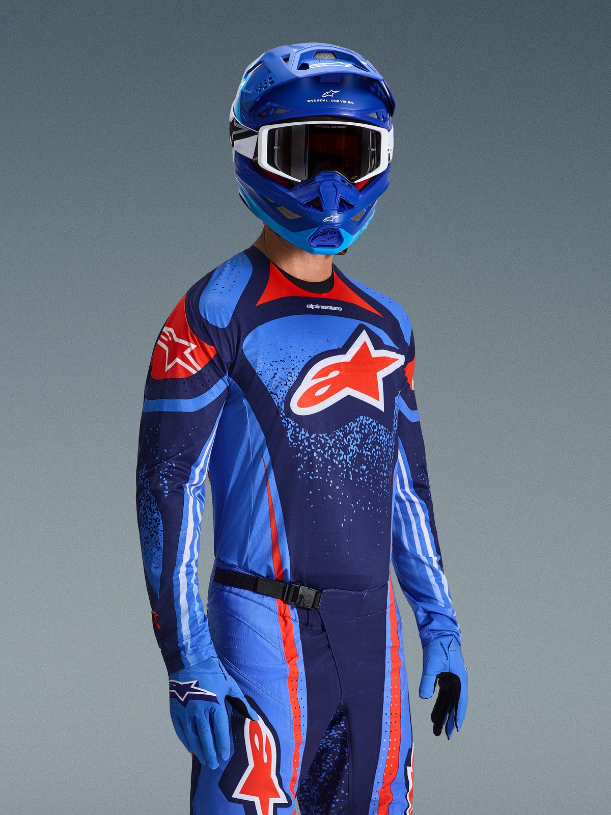 Piloto de motocross a usar a Camisola Alpinestars 2026 Techstar Nomur, equipamento de corrida em azul marinho, laranja e azul UCLA, com um grande logótipo Astars laranja, capacete e luvas azuis a condizer, conjunto de competição off-road atlético