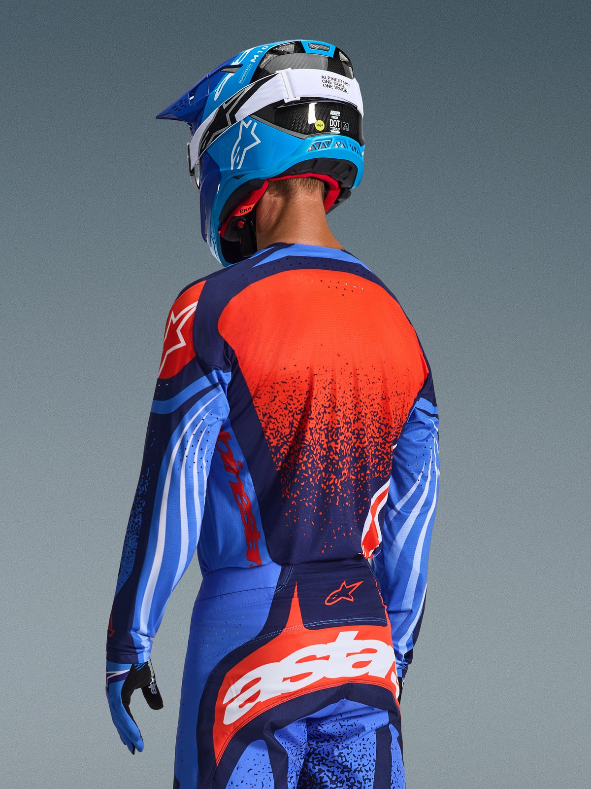Piloto de motocross visto de costas a usar a camisola Alpinestars 2026 Techstar Nomur e calças a condizer, em azul-escuro e azul UCLA com detalhes em laranja vibrante, com um grande grafismo de estrela e ventilação cortada a laser na camisola, combinado com um capacete azul e branco para um kit de competição todo-o-terreno completo