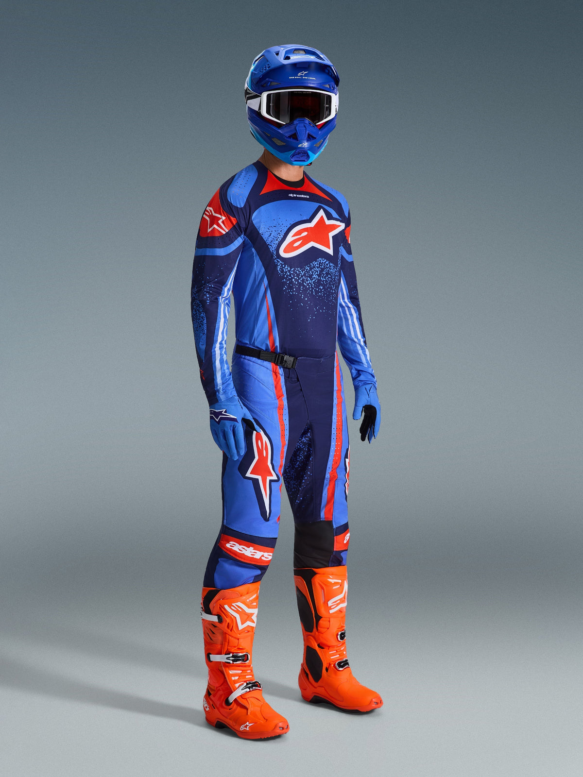 Piloto de motocross vestido com equipamento de corrida completo, camisola Techstar Nomur em azul e azul marinho escuro com logótipo Alpinestars laranja, calças a condizer com detalhes laranja, capacete azul com óculos brancos, botas cor de laranja vibrante e luvas azuis, conjunto coordenado para corridas off-road