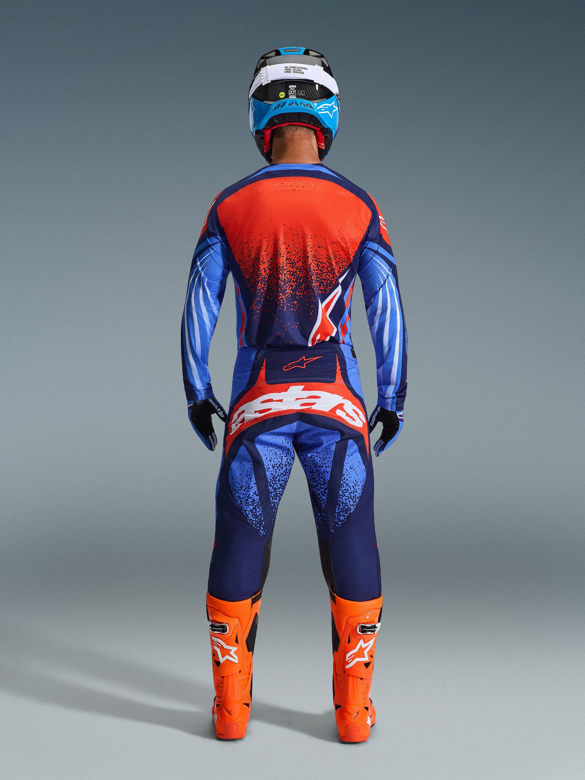 Piloto de motocross visto de trás a usar o conjunto de equipamento Alpinestars 2026 Techstar Nomur em Azul Marinho Escuro, Laranja e Azul UCLA, camisola com as costas em laranja salpicado, calças a condizer com logótipo branco em destaque no assento, botas laranja vibrante e capacete azul e branco, kit de corrida para todo-o-terreno coordenado.