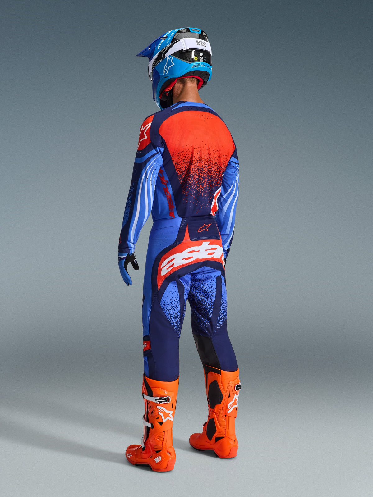 Piloto de motocross visto de costas a usar a camisola Alpinestars 2026 Techstar Nomur em azul-escuro, laranja e UCLA Blue, combinada com calças a condizer com logótipo branco em destaque, botas laranja vibrante e capacete azul e branco, kit completo de competição off-road.