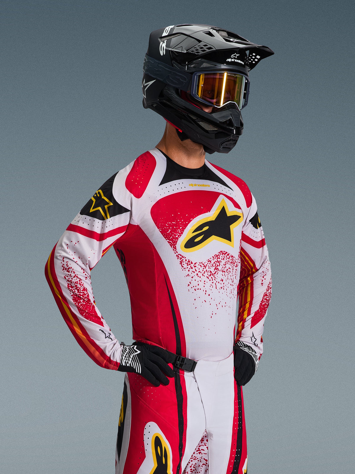 Piloto de motocross a usar o conjunto de equipamento Alpinestars 2026 Techstar Nomur, camisola branca e vermelha com pormenores pretos e logótipo amarelo, calças a condizer, capacete preto com óculos de lentes douradas, luvas pretas e brancas, ajuste atlético de competição para performance em todo-o-terreno